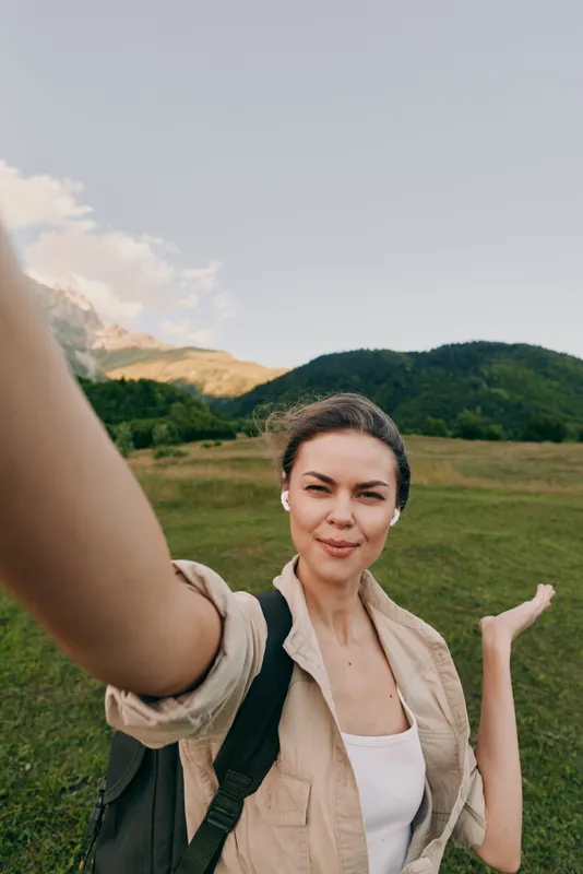 Woman takes a casual outdoor selfie in a green countryside field with a backpack, smiling under a bright sky with distant hills during a hike beside nature