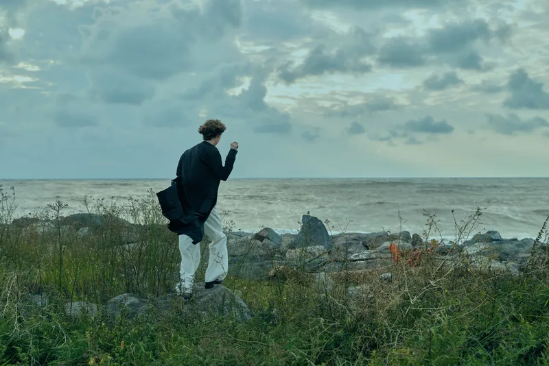 Man walking along a rocky coastline at dusk, jacket billowing, exploring the shore with ocean breeze and moody atmosphere, outdoor travel scene