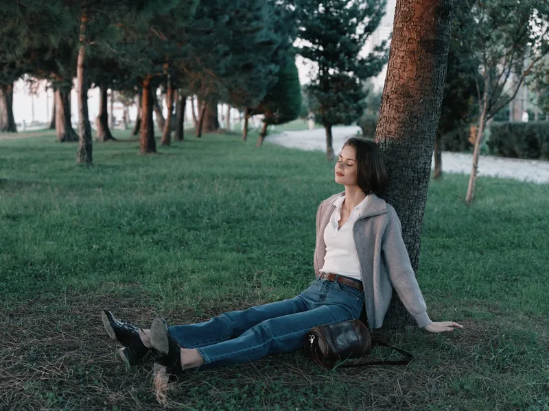 A relaxed professional woman sits against a tree in a park, smiling softly while resting after a walk. She wears casual business attire and comfortable shoes, creating a calm, reflective moment that