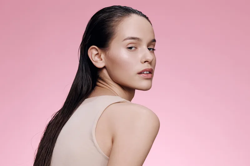 Elegant young woman with wet hair, posing against a soft pink background, conveying confidence and beauty in a minimalist fashion style