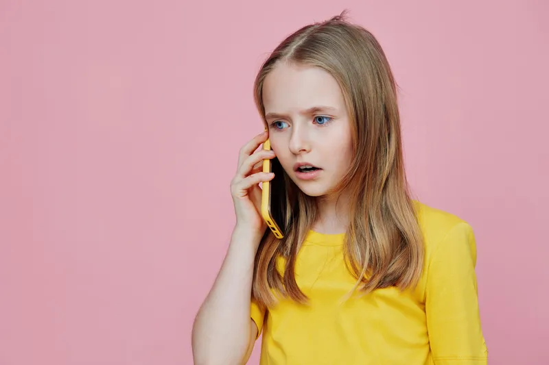 Concerned girl with long blonde hair talking on a yellow phone against a pink background, expressing emotions of surprise and worry in a vibrant summer outfit