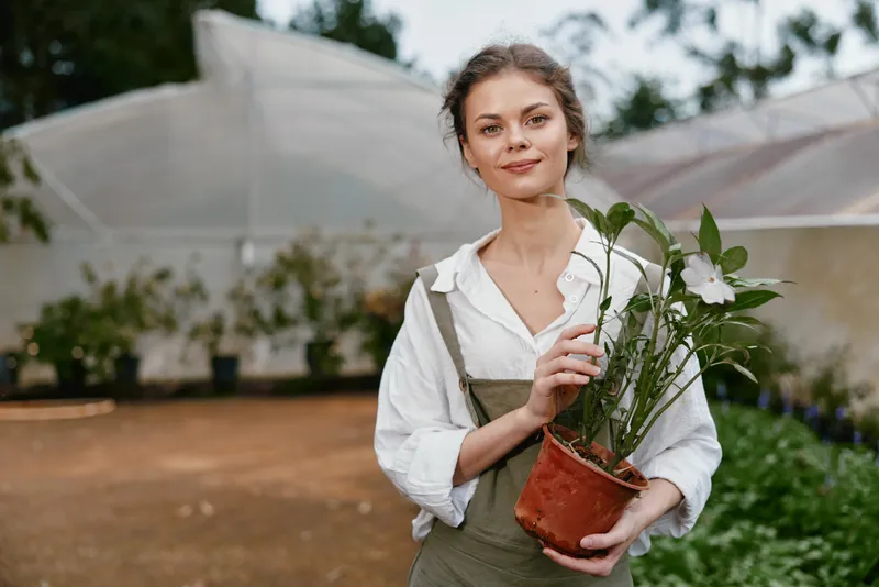 Young woman holding a potted plant in a greenhouse, displaying joy and passion for gardening with natural light and greenery around