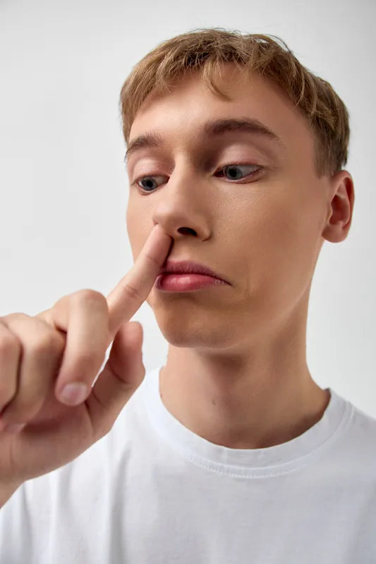 Closeup of a casual young man in a white tshirt pressing his finger to a nostril in a humorous, candid moment, minimal indoor light, simple background, and a focused, self aware expression.