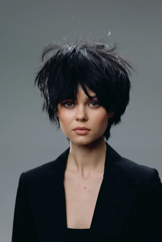 Young woman with short black hair and serious expression wearing a black jacket against gray background