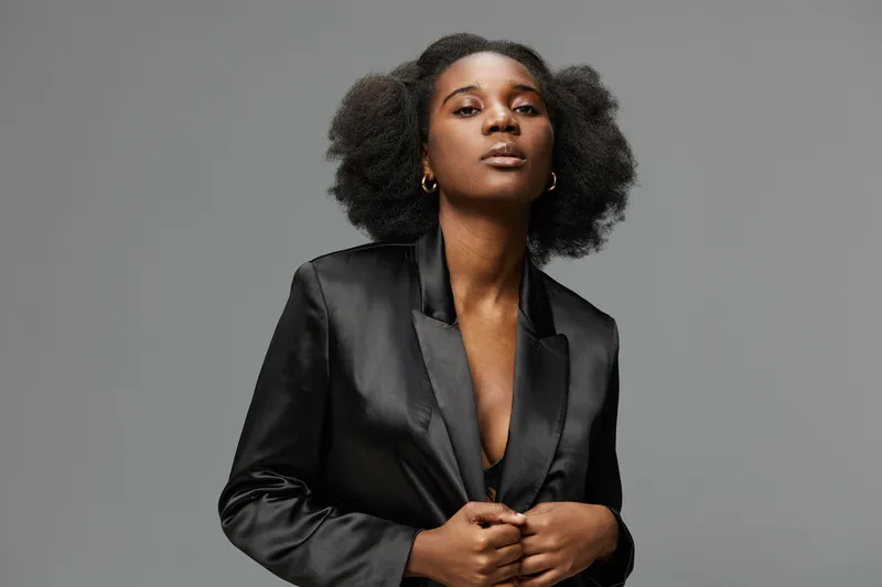 Black woman in a stylish black blazer displaying confidence with a natural hairstyle against a minimalist grey background