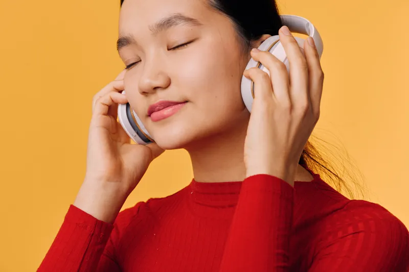Young woman in red sweater enjoying music with headphones on against soft yellow background with closed eyes and relaxed face for leisure and entertainment.
