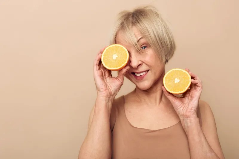 Smiling mature woman with short blonde hair holds two halves of orange, one covering eye. Beige background and casual clothing create fresh, healthy lifestyle and natural beauty concept.