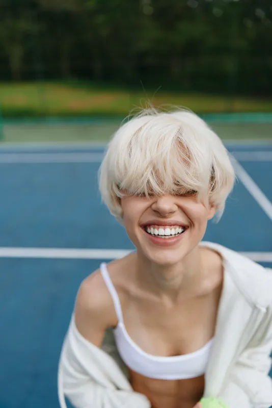 Young woman with short blonde hair smiling joyfully on a tennis court, wearing sporty attire, conveying a sense of health and fitness in a vibrant outdoor setting