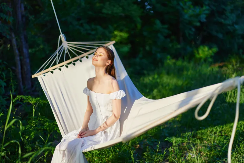 Young woman in a white dress relaxing in a hammock, enjoying nature during summer, surrounded by greenery, evoking tranquility and peace