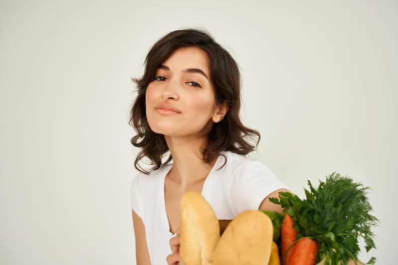 Жизнерадостная женщина с пакетом продуктов из супермаркета на светлом фоне
