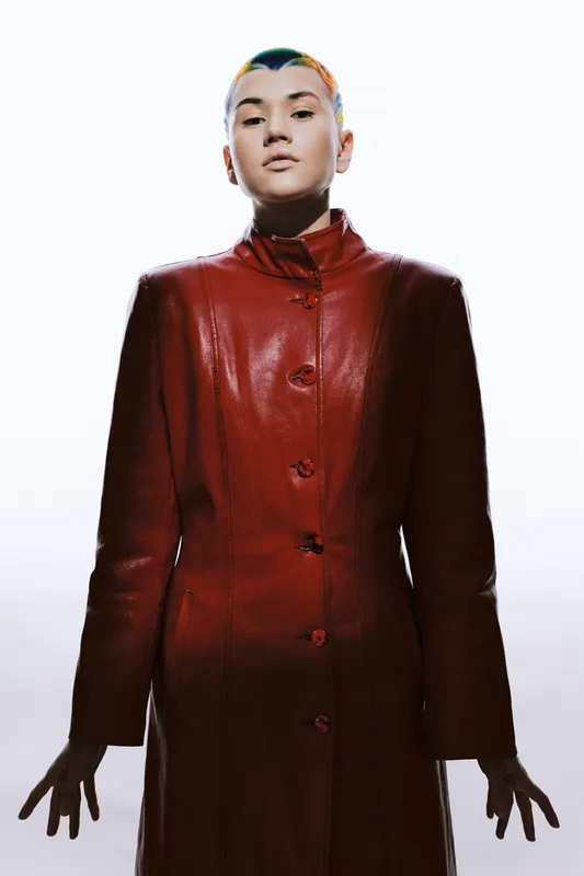 Young woman with short colorful hair wearing a long red leather coat standing confidently against white background with dramatic lighting and modern style.