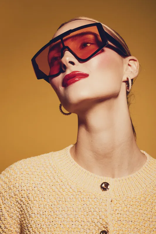 Stylish woman wearing oversized red sunglasses and a textured yellow sweater poses confidently against a warm yellow background. Concept of fashion, elegance, and modern style.
