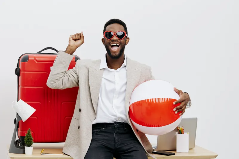 Excited man with beard wearing sunglasses, beige blazer and white shirt celebrating summer vacation at home, holding a striped beach ball, with a red suitcase in the background, studio shot, lifestyle