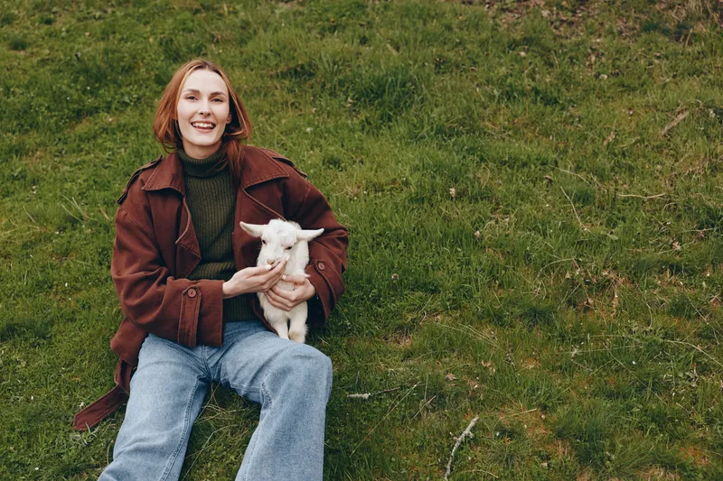 Smiling woman with goat kid sitting on grass in a natural outdoor setting, casual autumn outfit, joyful moment with animal, lifestyle scene, warm daylight, relaxed mood, connection with nature.