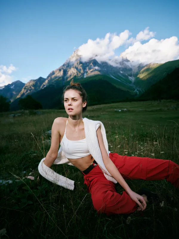 Outdoor mountains field model fashion casual athletic landscape sunlight nature, model seated in a grassy meadow wearing white crop top and red pants with towel draped over shoulder, bright lighting