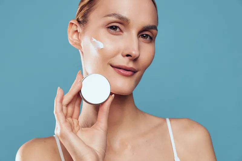 Young woman applies moisturizing cream on her face holds cosmetic container looks at camera with gentle smile isolated on blue background skincare beauty concept.