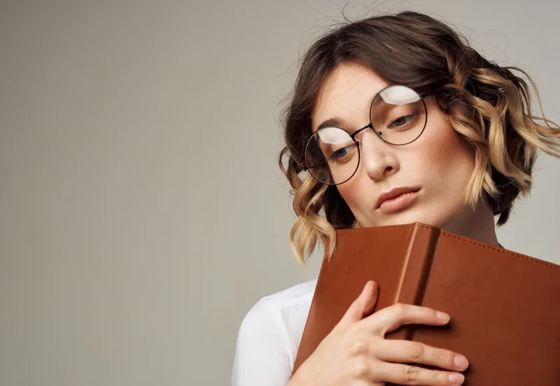 Романтичная женщина в очках на сером фоне с коричневой книгой в руках