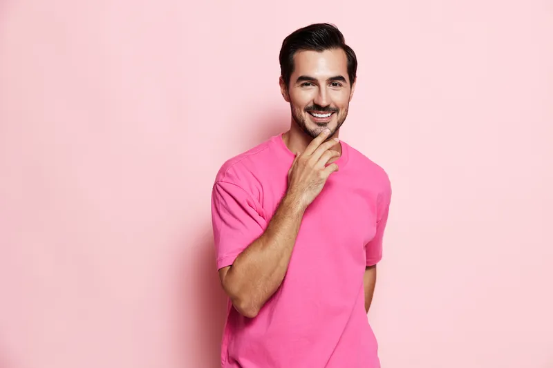Smartly dressed smiling man with short dark hair wearing a vibrant pink t-shirt poses confidently against a soft pink background. Positive vibe and lifestyle concept.