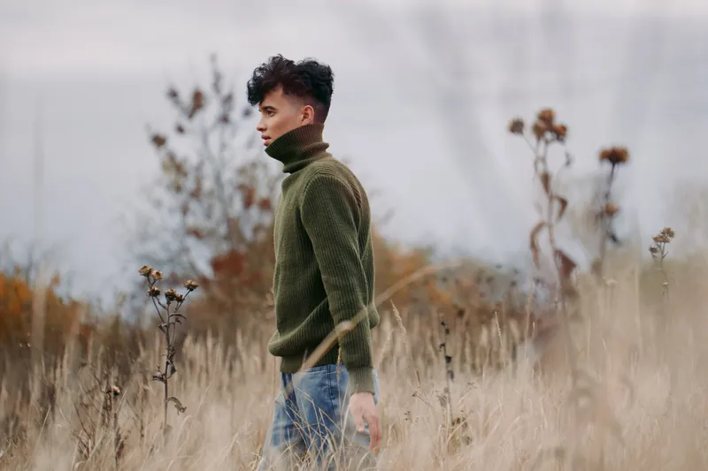 Young man walks through tall dry grass in a rustic meadow wearing a green turtleneck, captured in candid composition that highlights authenticity, credibility, natural texture and quiet, real emotion.