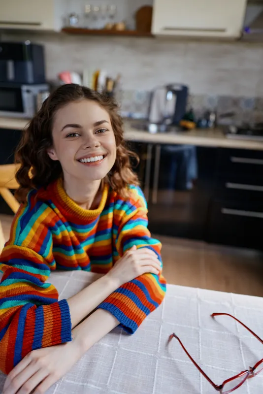 Smiling woman with curly hair wears a colorful striped sweater and sits at a kitchen table, relaxed and casual, highlighting a cozy home lifestyle with warm lighting and everyday comfort.