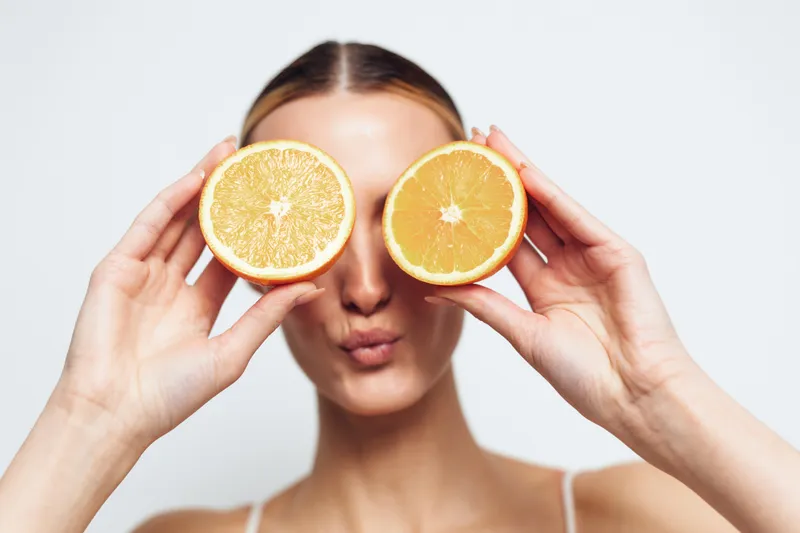 A young woman holds two halves of an orange in front of her eyes while puckering her lips. The bright studio background emphasizes the fresh fruit and playful mood.