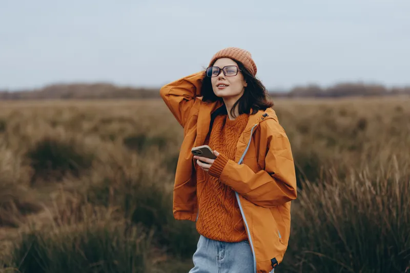 Outdoor woman lifestyle portrait in an autumn field, she wears a beanie, glasses, and a warm coat, one hand adjusting the collar, calm smile, soft daylight, nature background, gentle breeze