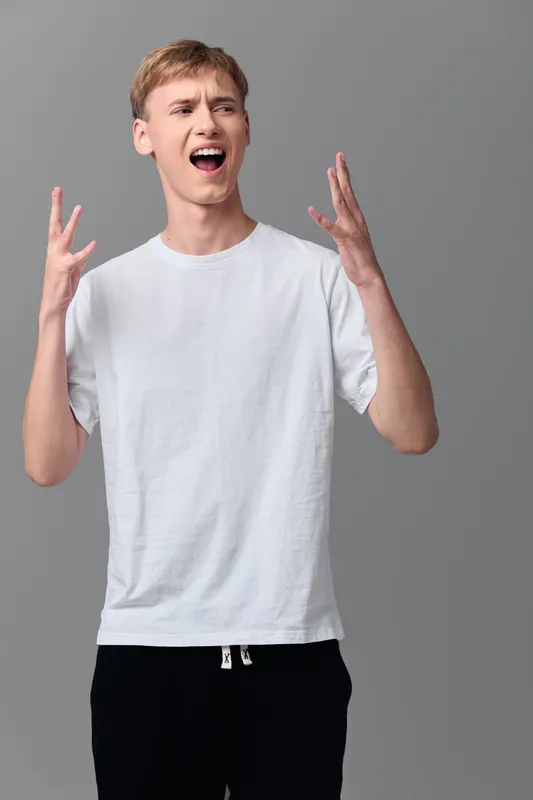 Shouting man in a white t shirt raises his hands with an open mouth, expressing surprise in a neutral studio for lifestyle, reaction, drama and energetic moments