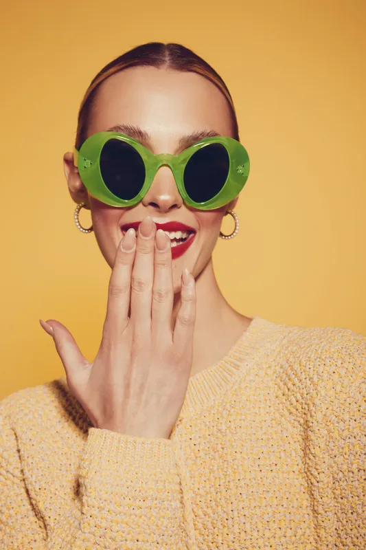 Young woman in green sunglasses and yellow sweater covers mouth with hand, smiling against yellow background. Concept of fashion, style, and playful mood.