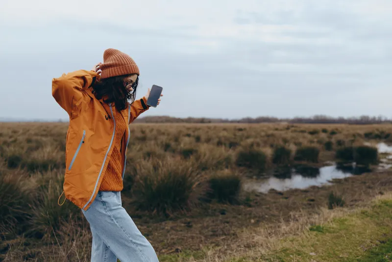 Woman lifestyle outdoors, nature field walk holding phone, wearing jacket and beanie, by small river in countryside, cloudy morning light, calm mood, travel and lifestyle concept.
