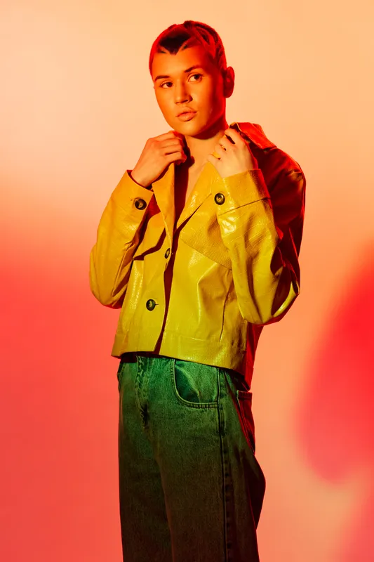 Young woman wearing a yellow jacket and green pants poses confidently against vibrant orange and red gradient background under studio lighting.