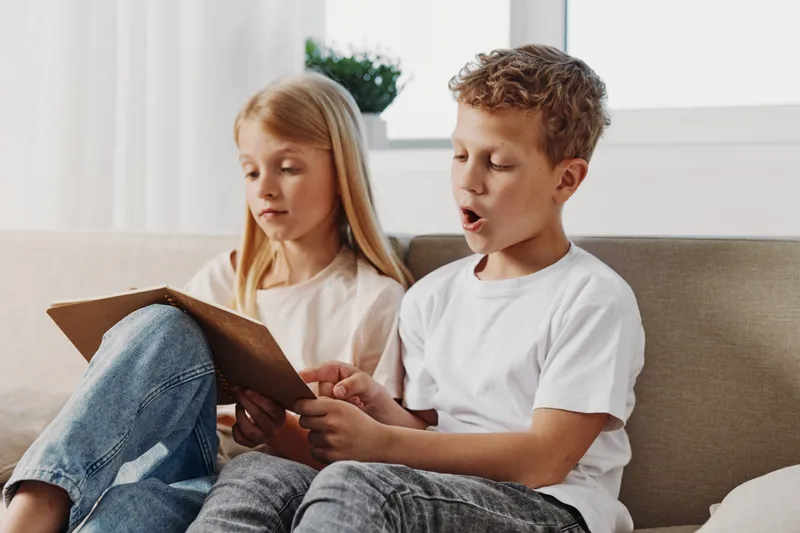 Two children sitting on a sofa reading a book together at home, boy enthusiastically narrates while girl listens attentively in a cozy living room setting.