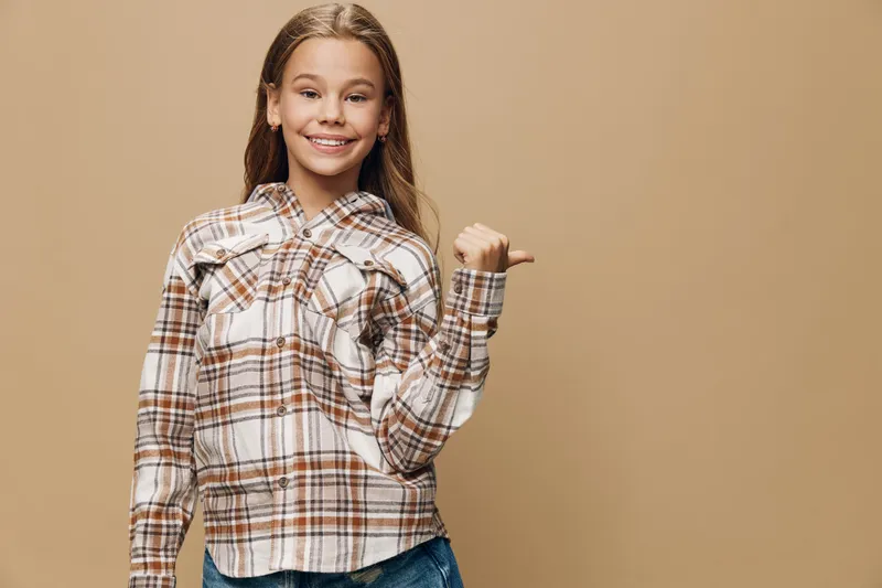Girl smiling and posing with thumb pointing sideways in casual plaid shirt and jeans, standing against beige background with relaxed and confident expression.