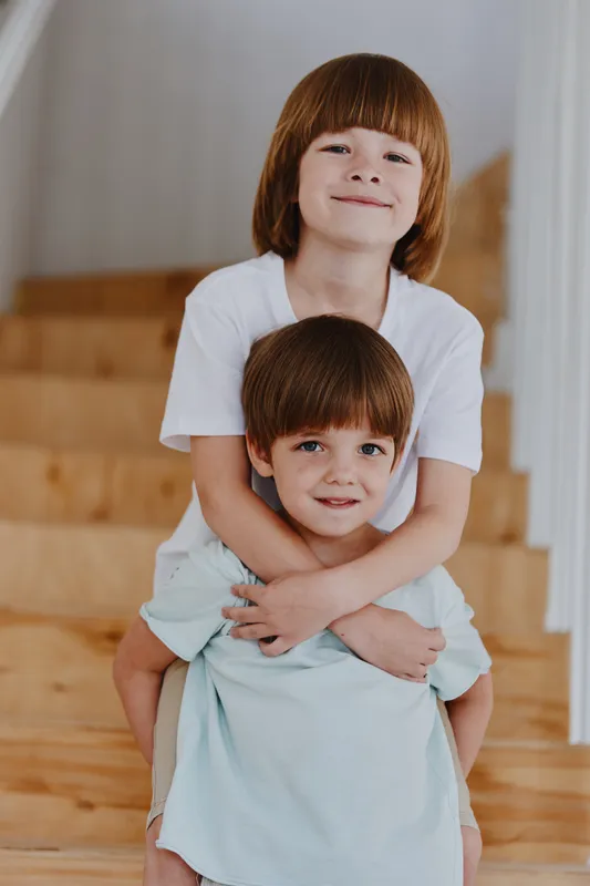 Two cheerful boys embracing each other on a wooden staircase, showcasing a joyful sibling bond, lively expressions and casual outfits.