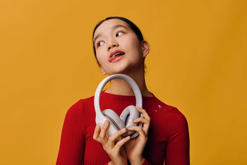 Young woman holding headphones in hands looking sideways with thoughtful expression on orange background. Casual female, fashion, technology, music accessory and lifestyle concept.