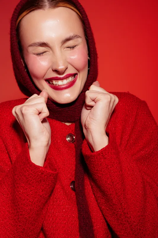 Happy woman in red wool coat and headscarf smiles with closed eyes and clenched fists against red background. Concept of joy, excitement, winter fashion, and positive emotions.