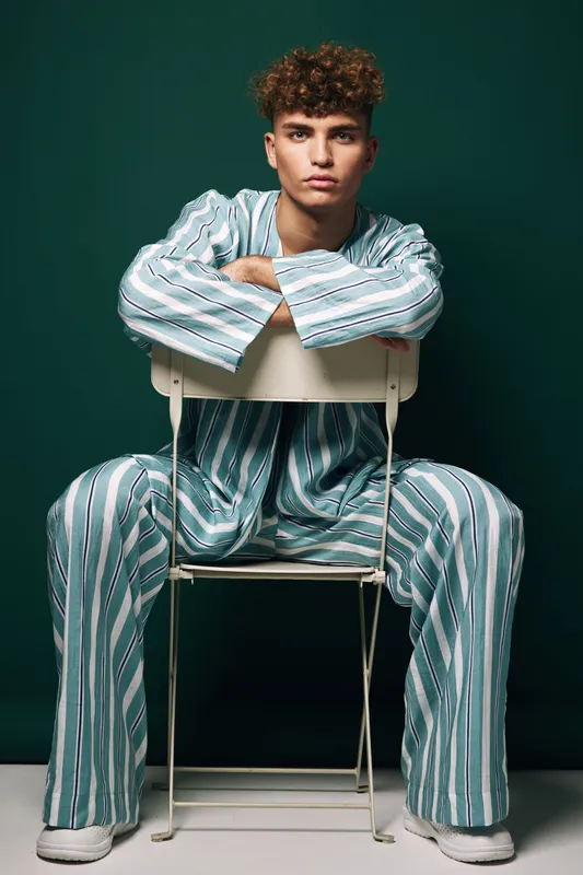 Young man with curly hair wearing striped pajamas sitting on white chair against dark green background looking serious and confident in studio portrait.