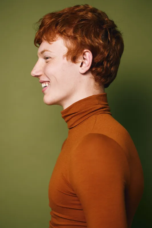Young man with red hair wearing brown turtleneck sweater smiling in profile on green background. Portrait of teenager with curly hairstyle looking happy and relaxed indoors.