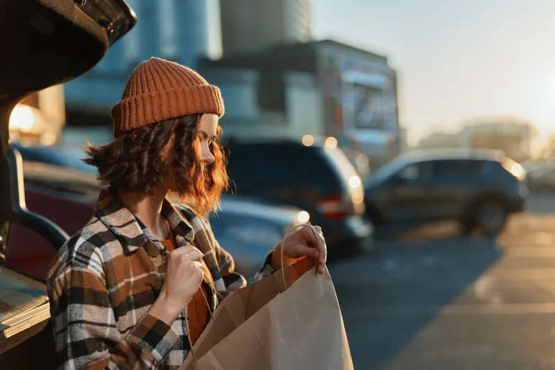 Woman car parking paperbag shopping goldenhour candid authenticity lifestyle urban scene: young woman in beanie and plaid jacket opening a paper bag at trunk, warm backlit moment of mindful living.