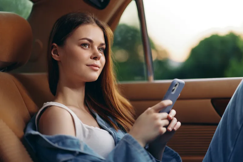 Young woman with long hair sitting in a car and using a smartphone, casual outfit, relaxed expression, soft natural lighting, summer vibes.