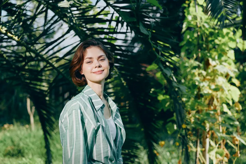 Portrait of a smiling woman standing in a sunlit garden with lush greenery and dappled light, conveying natural beauty and relaxed charm.