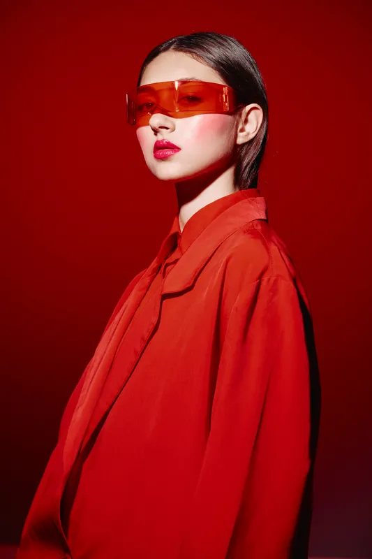 Fashionable young woman with sleek brown hair wearing oversized orange sunglasses and a trendy red jacket on a vibrant red background. Bold makeup highlights lips.