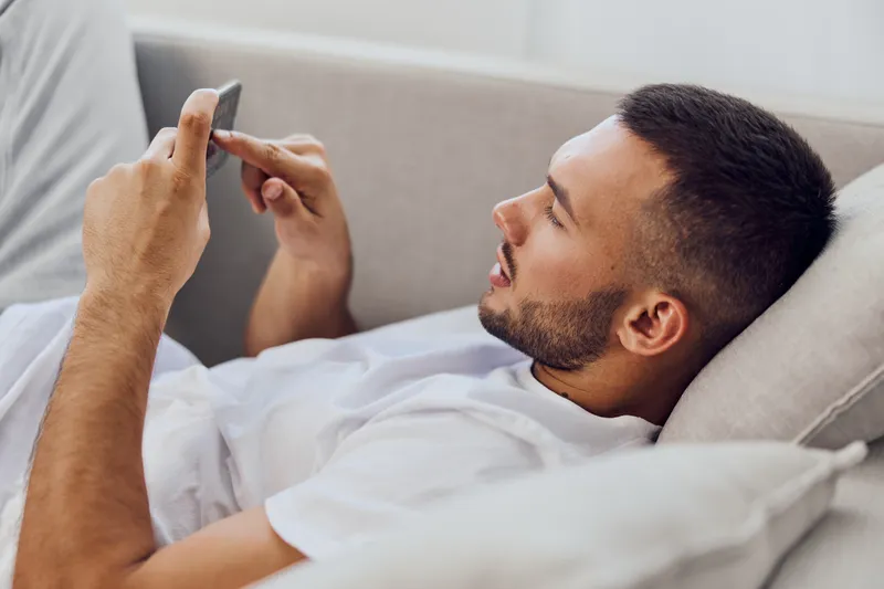 Relaxed man using smartphone while lounging on a cozy sofa, expressing leisure and modern lifestyle in a bright, inviting living room setting.