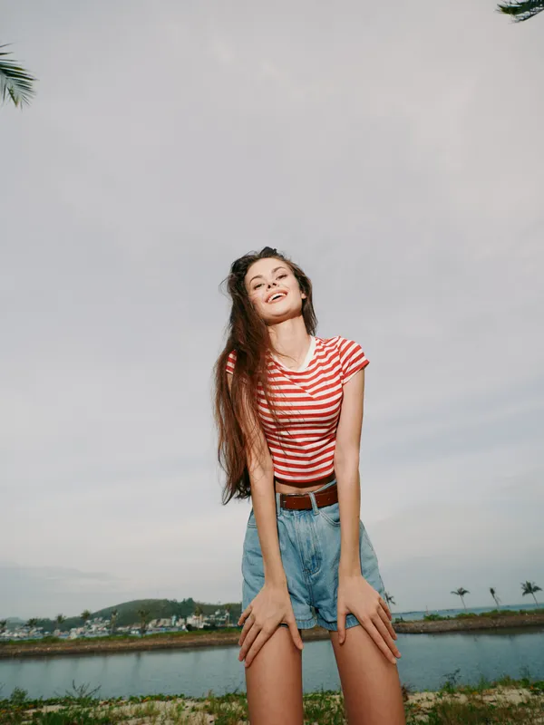 Young woman stands outdoors near a calm lake, wearing a red striped top and denim shorts, smiling with sunlit hair flowing, creating a carefree summertime portrait against a clear sky and distant