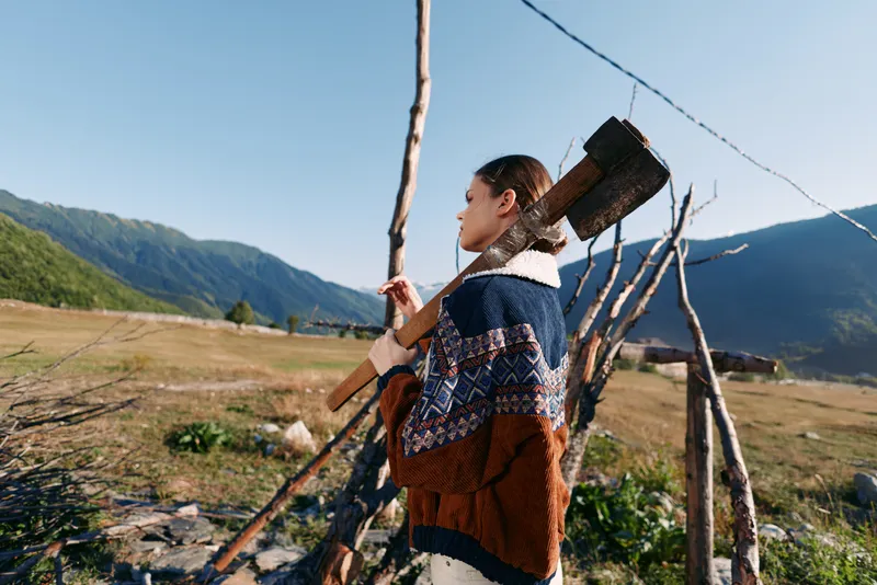 Woman with axe by fence in mountains field, wearing patterned sweater in rural wood landscape, carrying firewood over shoulder for outdoor countryside portrait and rustic lifestyle.