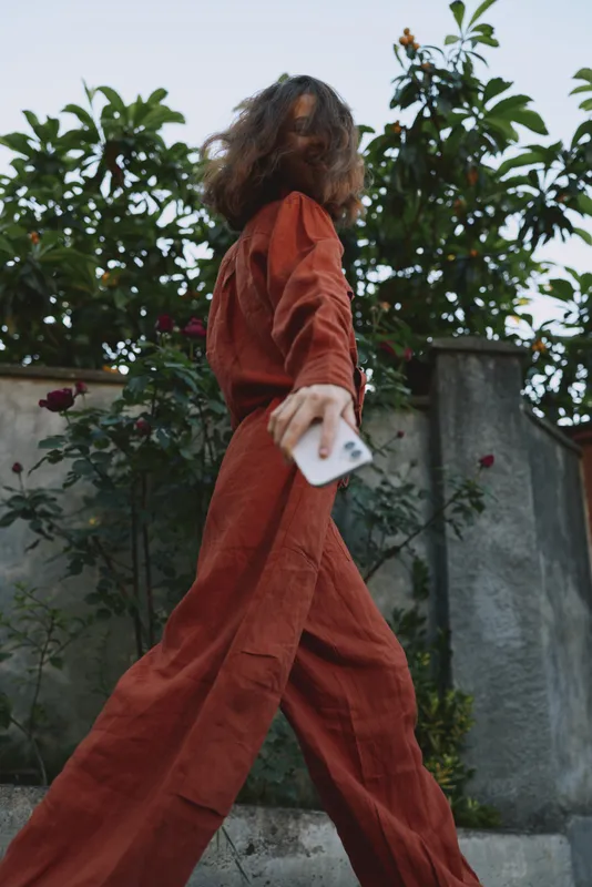 Woman in a rust red outfit walks outdoors with movement and stride, a candid lifestyle fashion shot emphasizing outdoor style, wind in fabric, and confident motion. Natural light enhances mood