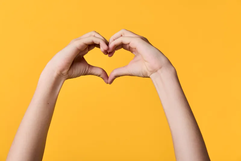 Hands form a heart gesture against a bright orange background, conveying love, affection and friendship with a warm, positive mood and uplifting message perfect for campaigns about unity, care, or