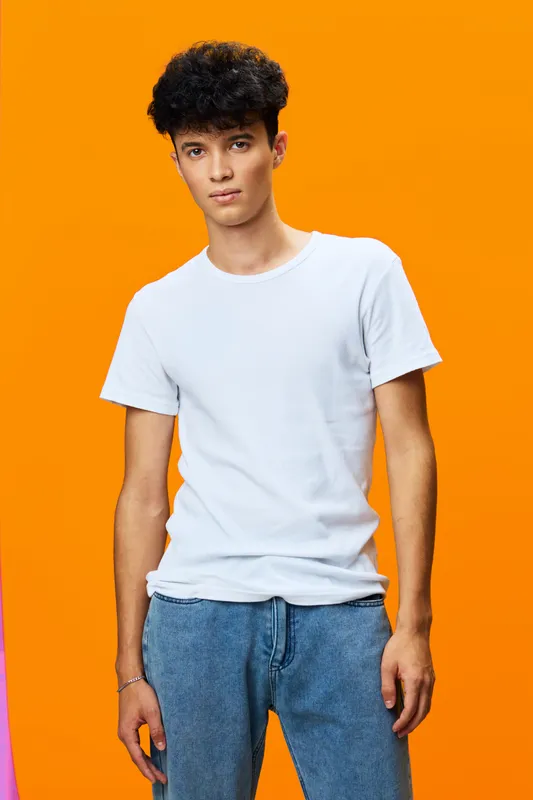young man with curly hair wearing a white t-shirt and blue jeans standing against a bright orange background with a serious expression