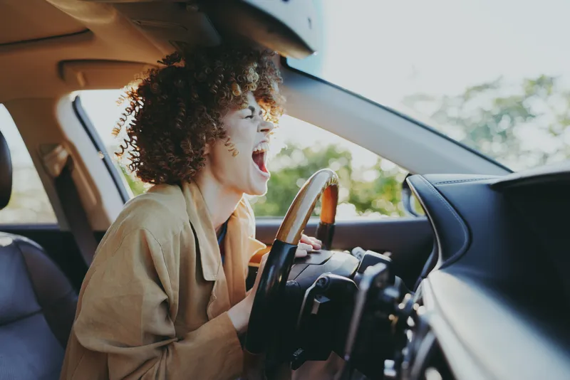 Young woman with curly hair drives in the car, shouting with excitement as the warm interior and casual beige cardigan convey a carefree road trip moment full of energy.
