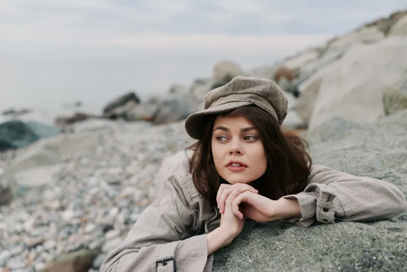 Young woman in casual vintage clothing lies on a rock at the rocky beach, gazing thoughtfully to the side. Overcast sky creates calm and reflective mood in nature.