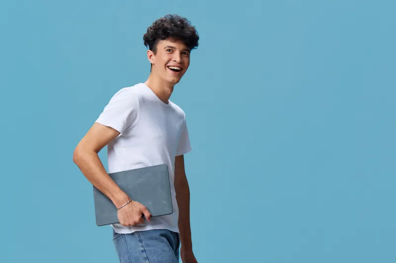 smiling man holding laptop in casual white t-shirt and jeans, standing against blue background with joyful expression for technology or work concept